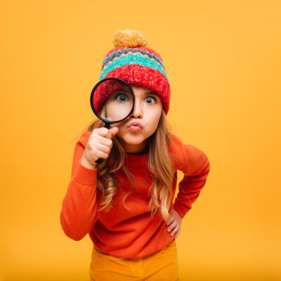 Joyful Young girl in sweater and hat looking at the camera with magnifier over orange background